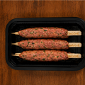 Three uncooked kebabs on sticks in a black plastic tray on a wooden surface.